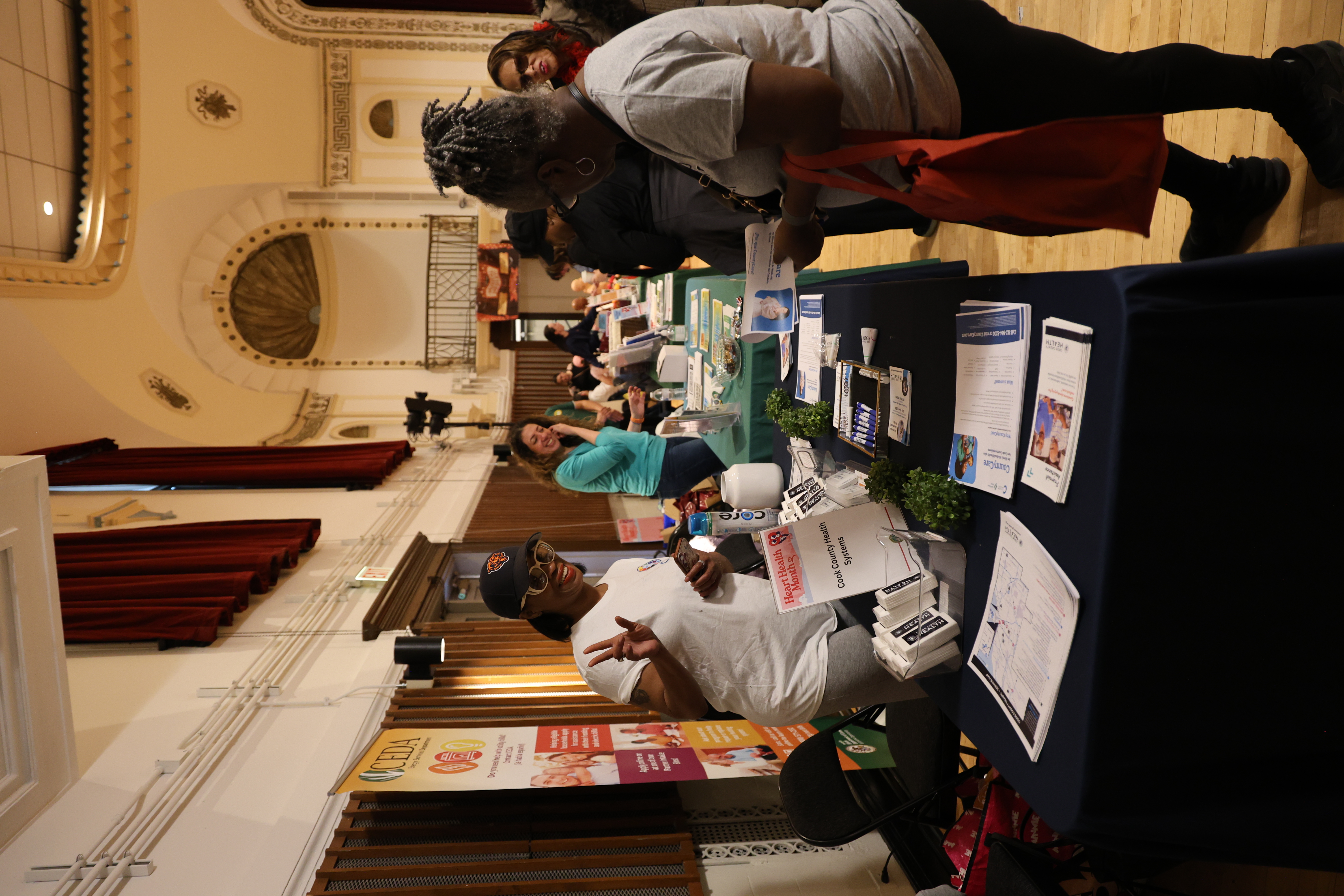Smiling woman in a cap makes a peace sign at a health fair booth with pamphlets; ornate hall background.